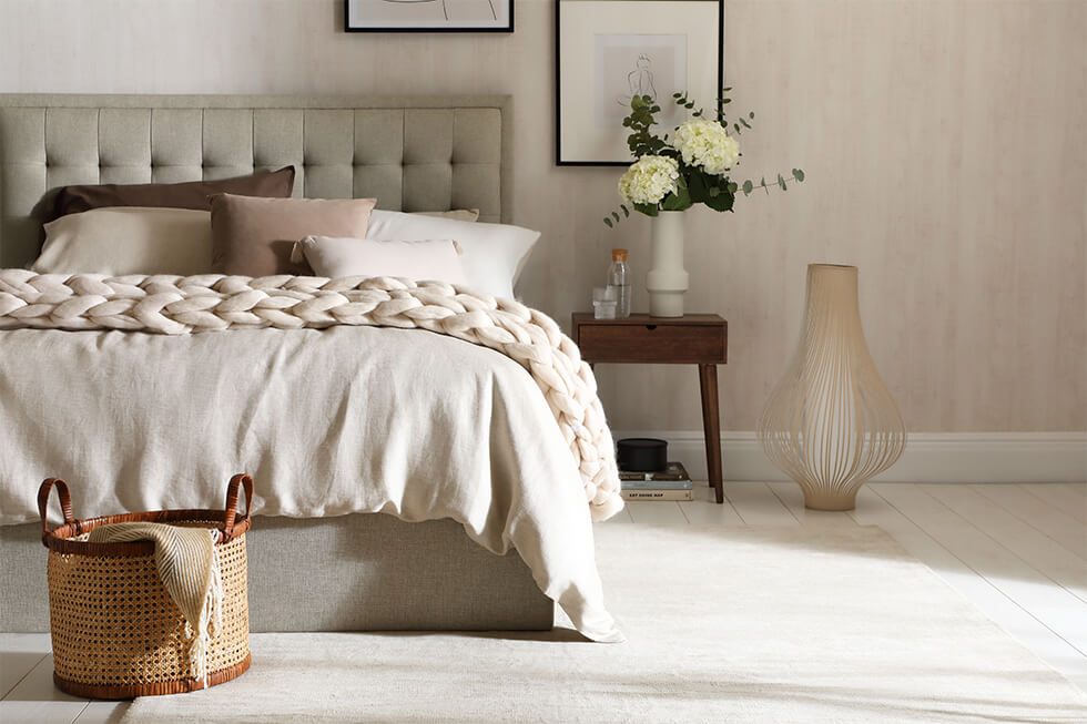 Cream and neutral bedroom with a double bed, luxurious pillows and throw, laundry basket, a side table with white roses, a vase, and two hanging paintings