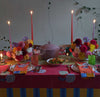 Dining table set with a pink floral tablecloth with a yellow and blue trim, white plates, orange napkins, colourful glasses, tapered candles, two bouquets of brightly coloured flowers, a lime-green table runner, and a pink ceramic pot in the centre