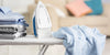 White ironing board with, from left to right, a pile of blue, white and grey clothes, an iron, and a blue and white striped shirt, on top