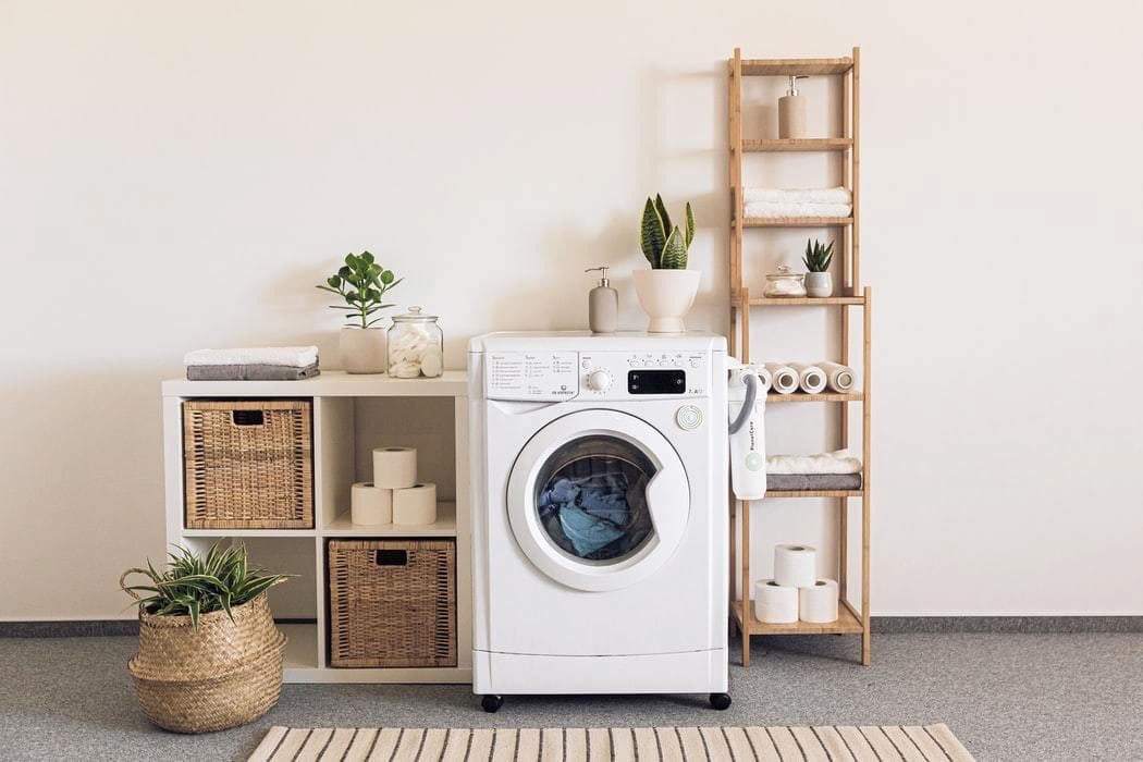From left to right, photo showing a basket with a plant inside, a shelf unit containing fitted baskets and tissue paper, a washing machine with a potted plant on top, and a ladder shelving unit holding towels, toilet rolls and linen