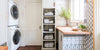 Modern luxe laundry room, from left to right: two double-stacked washing machines, towels in metal baskets in an open-storage system, counter with geometric tiles, green plants and window, patterned wallpaper