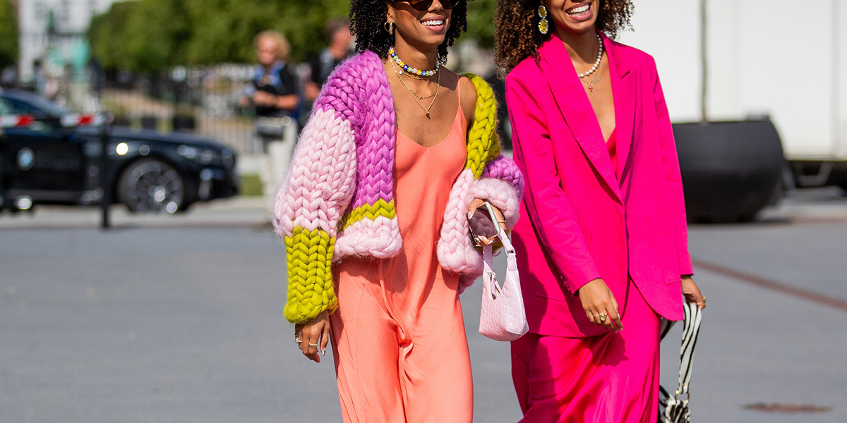 Photo of two women, one in a coral dress with a colour knitted cardigan over the top and the other in a bright pink suit, walking outside during Copenhagen Fashion Week