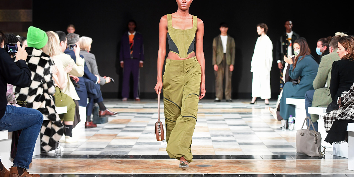 Model walking on a runway wearing a green and black corset top and green trousers made of natural materials
