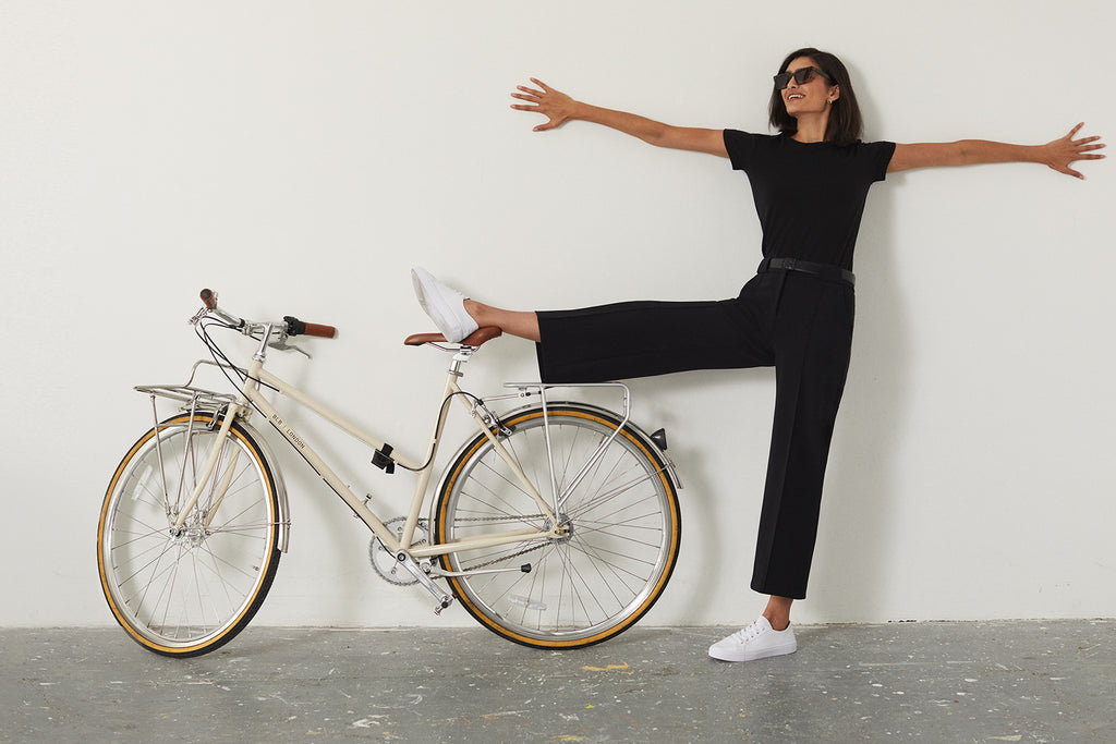 Dai model wearing black performance wear and white trainers lifting one leg over a bicycle with both arms outstretched