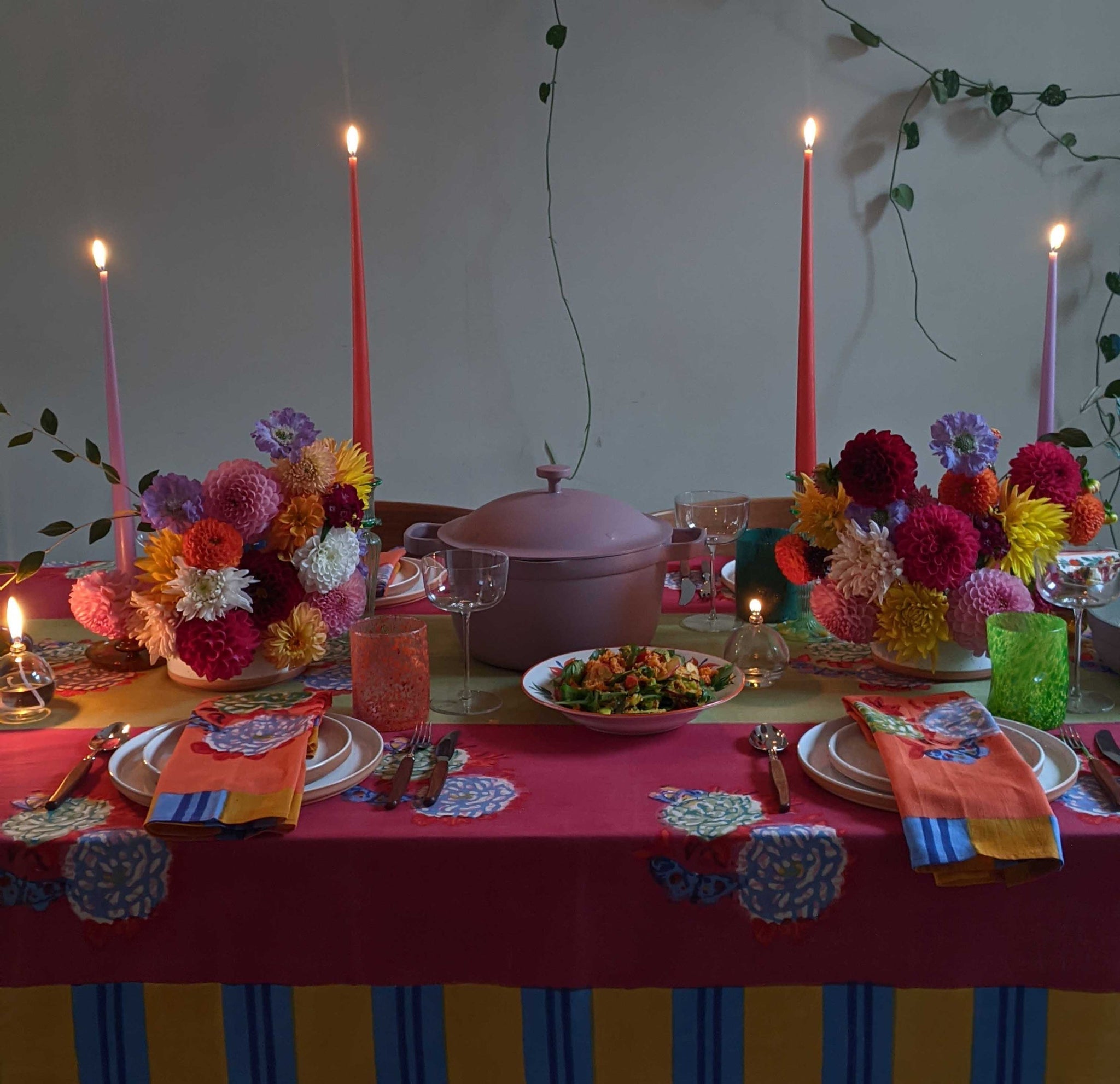 Dining table set with a pink floral tablecloth with a yellow and blue trim, white plates, orange napkins, colourful glasses, tapered candles, two bouquets of brightly coloured flowers, a lime-green table runner, and a pink ceramic pot in the centre 