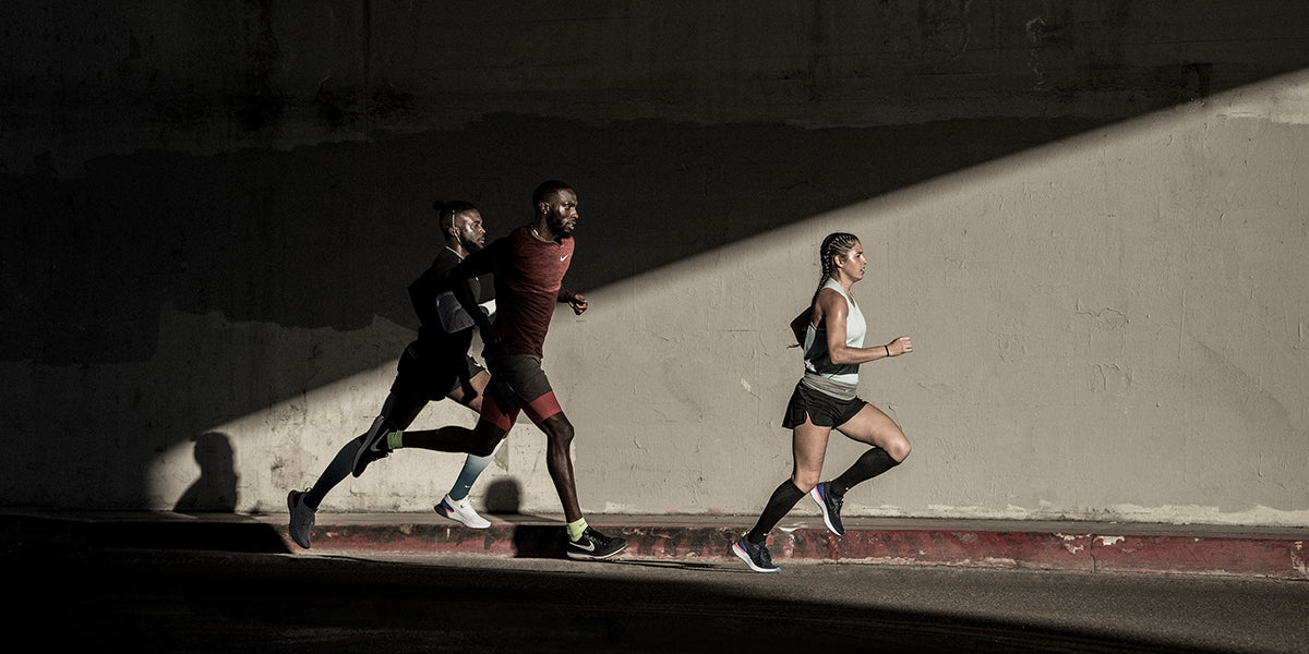 A woman and two men running outside on a street together in workout gear, woman is running in front. Background is a wall, half in shadow and half in sunlight.