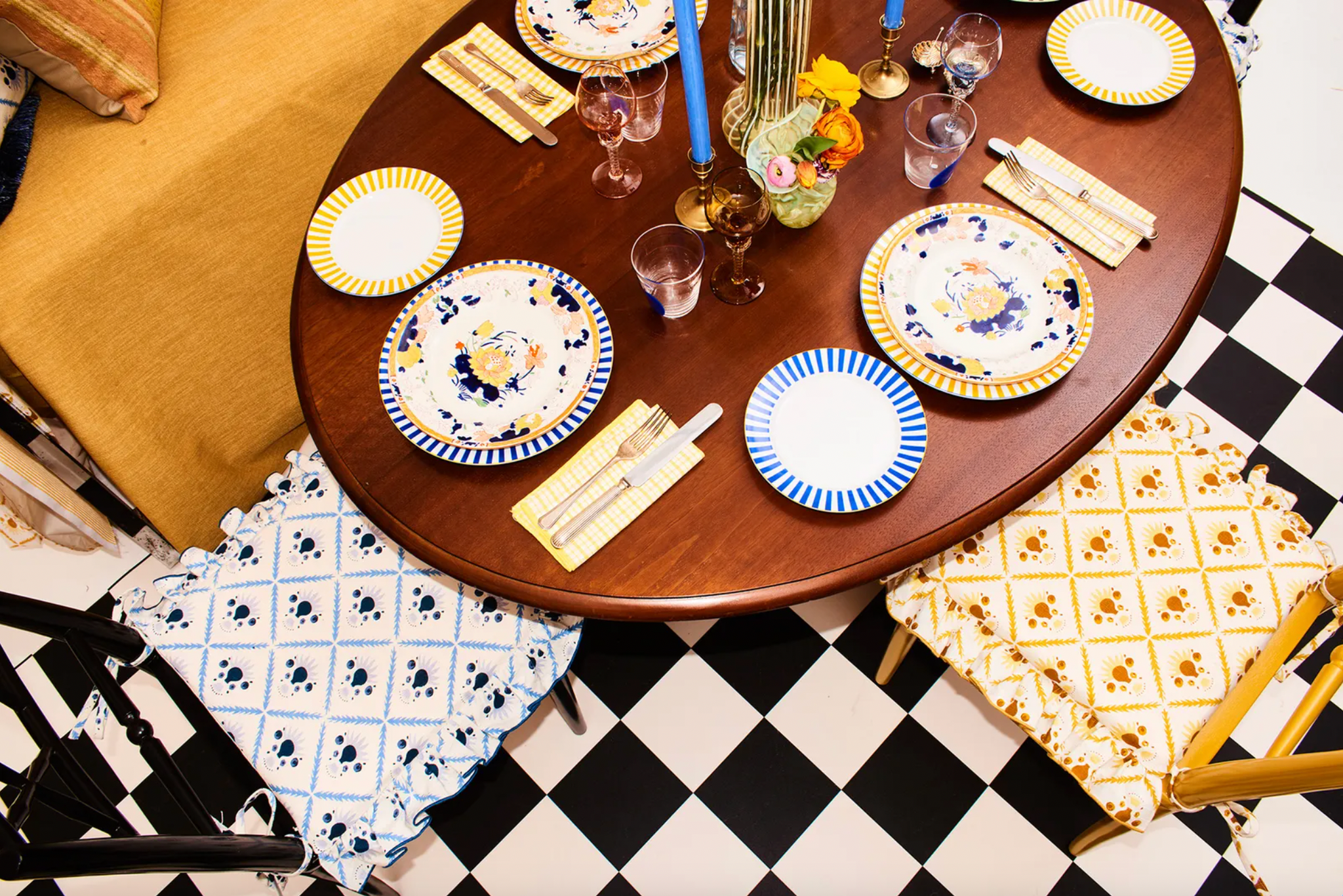 Round wooden dining table set with patterned crockery and blue tapered candles, with blue and yellow prints chairs and a chequered flooring