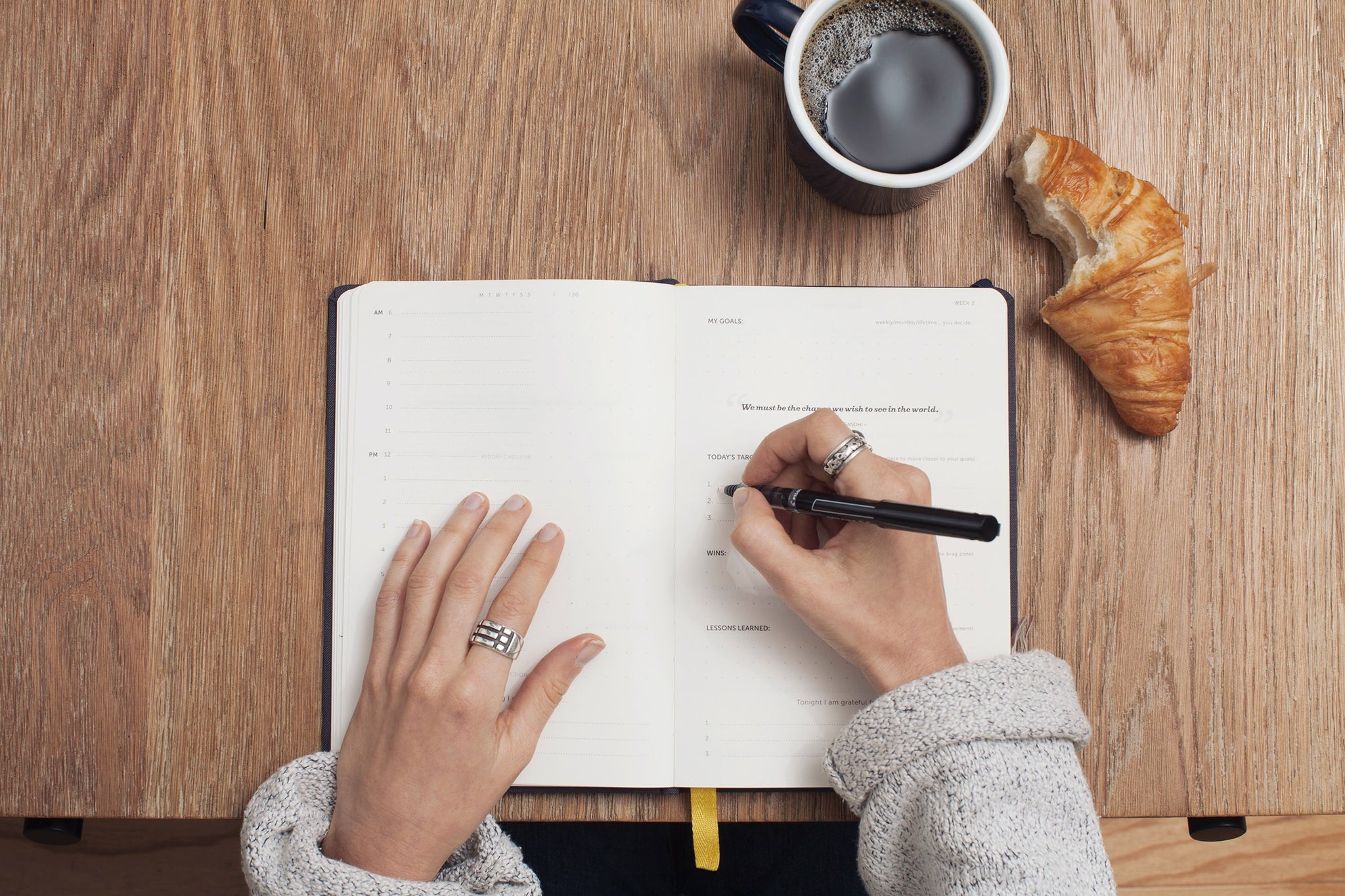 Woman writing in a diary/planner on a wooden table with a cup of coffee and a croissant next to her hands