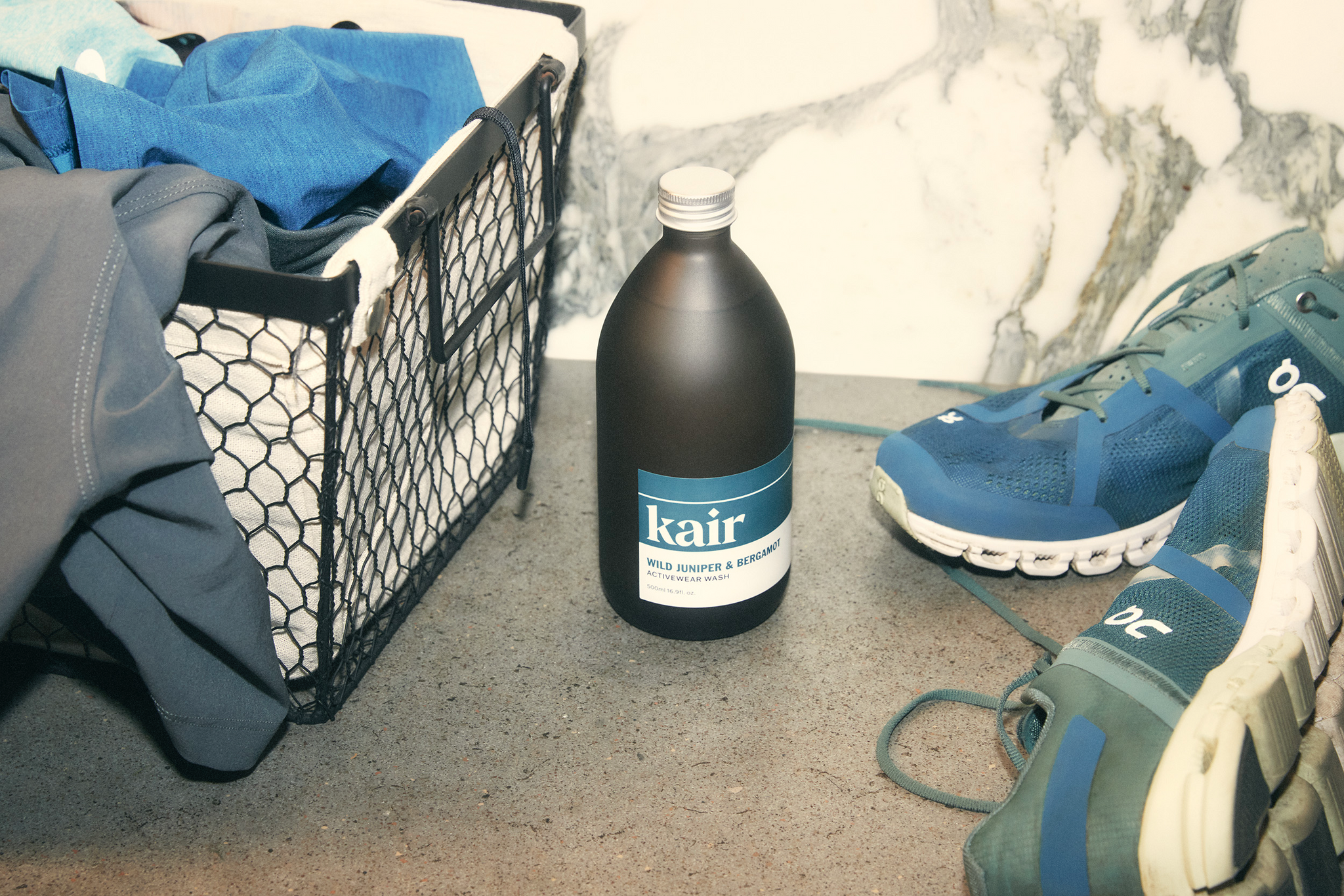 Photo of Kair's Activewear Wash bottle sitting between a laundry basket of dirty gym clothes and a pair of blue trainers