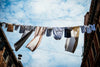 Photo looking up at a washing line between two buildings with a variety of clothes hanging and blowing in the wind against a cloudy blue sky